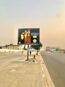 An urban street scene featuring a large billboard advertising a beverage. The billboard, set against a clear sky, displays two bottles and some promotional text in French. The street is lined with a sidewalk and a few pedestrians are walking. A lone tree stands near the billboard, and a car is seen driving down the road.