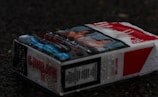 A wet cigarette pack lies on a dark surface, displaying health warnings and graphic images.