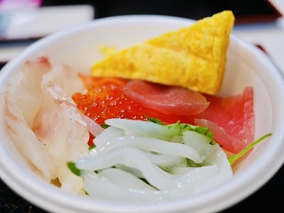 A bowl containing an assortment of Japanese sashimi on a bed of rice, featuring thinly sliced white fish, pink tuna, red salmon roe, translucent squid, and a triangular yellow piece of sweet omelette. Fresh greens are also visible.