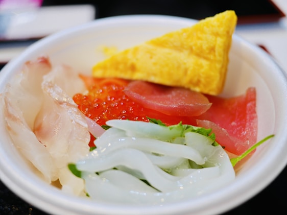 A bowl containing an assortment of Japanese sashimi on a bed of rice, featuring thinly sliced white fish, pink tuna, red salmon roe, translucent squid, and a triangular yellow piece of sweet omelette. Fresh greens are also visible.