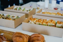 Several cardboard boxes filled with sandwiches and pastries are arranged on a table. The sandwiches, cut into portions, feature various bread types and fillings, including lettuce and other ingredients. In the foreground, croissants are visible on a wooden board, suggesting a casual or catered meal setting.
