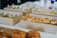 Several cardboard boxes filled with sandwiches and pastries are arranged on a table. The sandwiches, cut into portions, feature various bread types and fillings, including lettuce and other ingredients. In the foreground, croissants are visible on a wooden board, suggesting a casual or catered meal setting.