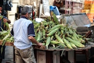 A man, seen from the back, is handling a large pile of corn on a wooden stall or table in a market setting. He is wearing a light blue shirt with dark sleeves and beige pants. The scene appears busy, with various items and structures in the background, including some metal and plastic items, highlighting the market atmosphere.