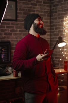 A bearded man wearing a black knit beanie and a burgundy shirt is standing indoors, holding a smartphone. He is smiling and appears relaxed. The background features a brick wall, string lights, and a desk with a lamp and a can of Coca-Cola.