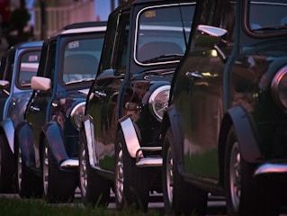 A row of classic cars parked closely together, with a focus on the shiny exteriors and rounded headlights. The cars have a vintage aesthetic and are aligned in a neat formation, likely in a residential area.
