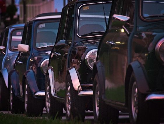 A row of classic cars parked closely together, with a focus on the shiny exteriors and rounded headlights. The cars have a vintage aesthetic and are aligned in a neat formation, likely in a residential area.