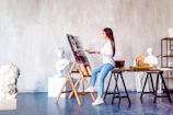 A woman is painting on a canvas set on an easel in an art studio. She is wearing a white shirt and jeans, holding a palette and brush. Surrounding her are several white plaster busts and sculptures. The studio has a minimalist and artistic feel, with a plain backdrop and wooden flooring.