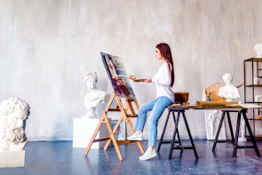 A woman is painting on a canvas set on an easel in an art studio. She is wearing a white shirt and jeans, holding a palette and brush. Surrounding her are several white plaster busts and sculptures. The studio has a minimalist and artistic feel, with a plain backdrop and wooden flooring.