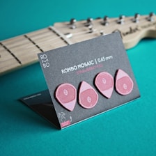 A set of four pink guitar picks displayed on a black card labeled 'ROMBO MOSAIC | 0.65 mm STRAWBERRY RED' stands on a turquoise surface. The background shows a blurred section of a wooden guitar neck with strings.