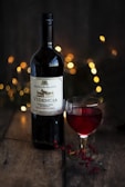 A bottle of red wine, labeled 'VIDENCIA', is set on a rustic wooden surface. Beside it is a glass of the same red wine, filled partially. The background is softly illuminated by warm, golden bokeh lights, creating a cozy and inviting atmosphere. Nearby, small red berries are scattered on the table, adding a touch of natural elegance.