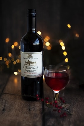 A bottle of red wine, labeled 'VIDENCIA', is set on a rustic wooden surface. Beside it is a glass of the same red wine, filled partially. The background is softly illuminated by warm, golden bokeh lights, creating a cozy and inviting atmosphere. Nearby, small red berries are scattered on the table, adding a touch of natural elegance.