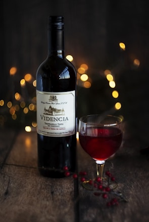 A bottle of red wine, labeled 'VIDENCIA', is set on a rustic wooden surface. Beside it is a glass of the same red wine, filled partially. The background is softly illuminated by warm, golden bokeh lights, creating a cozy and inviting atmosphere. Nearby, small red berries are scattered on the table, adding a touch of natural elegance.