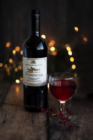 A bottle of red wine, labeled 'VIDENCIA', is set on a rustic wooden surface. Beside it is a glass of the same red wine, filled partially. The background is softly illuminated by warm, golden bokeh lights, creating a cozy and inviting atmosphere. Nearby, small red berries are scattered on the table, adding a touch of natural elegance.
