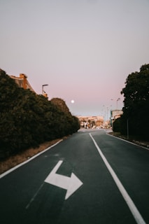 A road in an urban setting during early evening or dawn, characterized by a single white arrow painted on the asphalt, pointing to the left. The road is flanked by trees and leads towards a distant cityscape under a pale, pastel-colored sky with a visible moon.