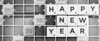 Letter tiles on a Scrabble board spell out the greeting 'Happy New Year.' The tiles are arranged horizontally in two rows with the words legible and positioned neatly among other tiles marked with 'double letter' and 'triple letter' indications.