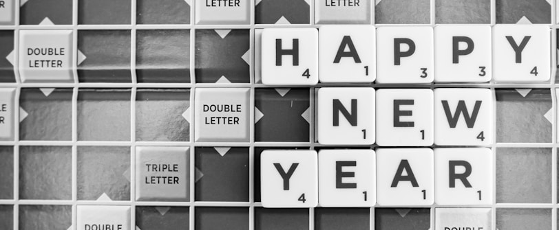 Letter tiles on a Scrabble board spell out the greeting 'Happy New Year.' The tiles are arranged horizontally in two rows with the words legible and positioned neatly among other tiles marked with 'double letter' and 'triple letter' indications.