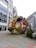 A large, colorful sculpture resembling a tangled mass of objects wrapped in thick rope is placed in an outdoor urban area. The sculpture includes various recognizable shapes like chairs, cubes, and bottles in vibrant colors. Surrounding it are modern office buildings with glass windows, creating a contrast between the art piece and its urban environment.