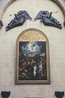 A large painting is framed and mounted on a stone wall, depicting numerous figures engaged in various dynamic postures, with some figures appearing to be ascending. The scene is highly detailed, showing expressions and movement, and is surrounded by ornate wooden carving. Above the painting, two sculptures of angels holding trumpets are mounted, appearing to be blowing them. The wall itself is made of large stone blocks.