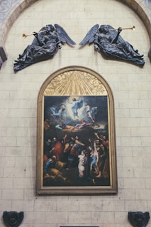 A large painting is framed and mounted on a stone wall, depicting numerous figures engaged in various dynamic postures, with some figures appearing to be ascending. The scene is highly detailed, showing expressions and movement, and is surrounded by ornate wooden carving. Above the painting, two sculptures of angels holding trumpets are mounted, appearing to be blowing them. The wall itself is made of large stone blocks.