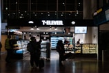 A 7-Eleven convenience store in an airport setting, featuring people with luggage browsing products inside. The store displays beverages, snacks, and has a counter with a salesperson. Bright lighting highlights the assortment of items.