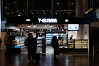 A 7-Eleven convenience store in an airport setting, featuring people with luggage browsing products inside. The store displays beverages, snacks, and has a counter with a salesperson. Bright lighting highlights the assortment of items.