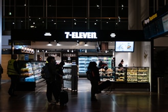 A 7-Eleven convenience store in an airport setting, featuring people with luggage browsing products inside. The store displays beverages, snacks, and has a counter with a salesperson. Bright lighting highlights the assortment of items.