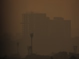 A hazy cityscape with silhouettes of tall buildings and towers barely visible through a thick layer of smog. The atmosphere is heavily polluted, giving the scene a murky orange-brown tint.