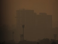 A hazy cityscape with silhouettes of tall buildings and towers barely visible through a thick layer of smog. The atmosphere is heavily polluted, giving the scene a murky orange-brown tint.