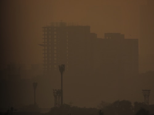 A hazy cityscape with silhouettes of tall buildings and towers barely visible through a thick layer of smog. The atmosphere is heavily polluted, giving the scene a murky orange-brown tint.