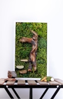 A vertical art piece featuring a textured combination of moss and various natural elements. A prominent driftwood-like structure is centrally embedded in a dense green moss background. Additional items include bark, smooth stone-like forms, and a few small branches or sticks. The whole piece is set within a white frame and rests on a wooden table, which also displays a pine cone, a small rock, and more twigs or branches.
