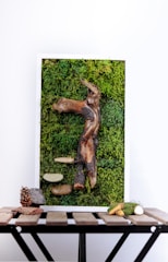 A vertical art piece featuring a textured combination of moss and various natural elements. A prominent driftwood-like structure is centrally embedded in a dense green moss background. Additional items include bark, smooth stone-like forms, and a few small branches or sticks. The whole piece is set within a white frame and rests on a wooden table, which also displays a pine cone, a small rock, and more twigs or branches.