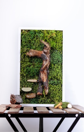 A vertical art piece featuring a textured combination of moss and various natural elements. A prominent driftwood-like structure is centrally embedded in a dense green moss background. Additional items include bark, smooth stone-like forms, and a few small branches or sticks. The whole piece is set within a white frame and rests on a wooden table, which also displays a pine cone, a small rock, and more twigs or branches.