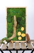 A vertical wooden frame contains a rich tapestry of green moss, with a prominent piece of textured driftwood and several sliced wooden discs arranged artistically. Below the frame, on a rustic wooden table, various natural objects like small branches, pine cones, and additional wood slices are scattered.