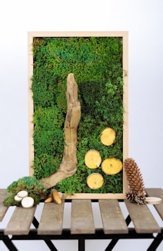 A vertical wooden frame contains a rich tapestry of green moss, with a prominent piece of textured driftwood and several sliced wooden discs arranged artistically. Below the frame, on a rustic wooden table, various natural objects like small branches, pine cones, and additional wood slices are scattered.