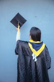 A person wearing a black graduation gown and a yellow and blue stole holds a graduation cap with a yellow tassel against a blue background.