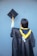 A person wearing a black graduation gown and a yellow and blue stole holds a graduation cap with a yellow tassel against a blue background.