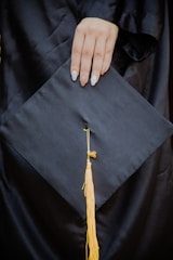 A hand with manicured nails rests on a black graduation cap, which features a yellow tassel hanging from the center.