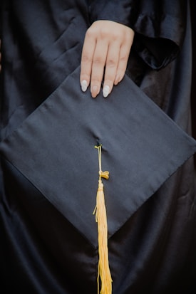 A hand with manicured nails rests on a black graduation cap, which features a yellow tassel hanging from the center.