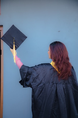 A person in a graduation gown is holding a graduation cap with a tassel. The background is a plain blue wall, and the person is facing away from the camera, showcasing long hair.