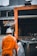 A sanitation worker wearing an orange uniform and hat stands next to a garbage truck and several trash bins. The truck and bins are situated in an urban area, possibly near shops or eateries, as a sign for a food place is visible in the background. The atmosphere appears overcast.