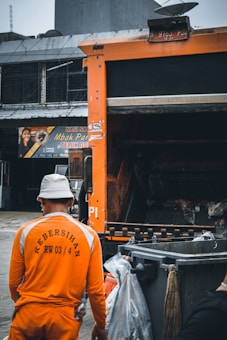 A sanitation worker wearing an orange uniform and hat stands next to a garbage truck and several trash bins. The truck and bins are situated in an urban area, possibly near shops or eateries, as a sign for a food place is visible in the background. The atmosphere appears overcast.