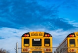Two yellow school buses are parked behind a chain-link fence under a bright blue sky. The rear views of the buses show prominently marked signs indicating they are school buses, with additional safety notices.