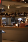 A kitchen area with a sign that says 'MAC & CHEESE' adorned with festive garlands. Two people are working behind the counter, one bent over concentrating on a task and the other standing. The setup includes a coffee machine and other kitchen tools on the counter. The atmosphere looks professional and busy.