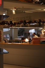A kitchen area with a sign that says 'MAC & CHEESE' adorned with festive garlands. Two people are working behind the counter, one bent over concentrating on a task and the other standing. The setup includes a coffee machine and other kitchen tools on the counter. The atmosphere looks professional and busy.