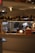 A kitchen area with a sign that says 'MAC & CHEESE' adorned with festive garlands. Two people are working behind the counter, one bent over concentrating on a task and the other standing. The setup includes a coffee machine and other kitchen tools on the counter. The atmosphere looks professional and busy.