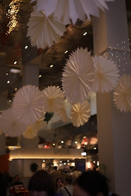 Large paper decorations resembling stars or fans hang from the ceiling in a dimly lit space. The area is busy, with people seated and walking around. The ceiling has mirrored surfaces and small recessed lights, adding to the ambiance. The overall atmosphere is warm and festive.