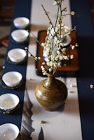 A rustic ceramic vase containing branches with white blossoms is placed on a table runner. Surrounding the vase are several white ceramic teacups, each with a unique blue pattern on a saucer. The setup is elegant and likely part of a traditional tea ceremony setting.