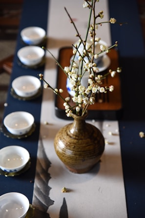 A rustic ceramic vase containing branches with white blossoms is placed on a table runner. Surrounding the vase are several white ceramic teacups, each with a unique blue pattern on a saucer. The setup is elegant and likely part of a traditional tea ceremony setting.
