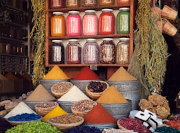 A vibrant market stall displaying various colorful spices in pyramidal heaps, metal containers, and woven baskets. Above the spices, there are jars filled with different dried herbs and ingredients neatly arranged on a wooden shelf. The scene is enhanced by hanging plants and an assortment of woven crafts, creating an atmospheric and lively setting.