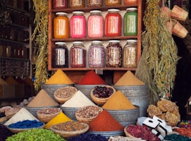 A vibrant market stall displaying various colorful spices in pyramidal heaps, metal containers, and woven baskets. Above the spices, there are jars filled with different dried herbs and ingredients neatly arranged on a wooden shelf. The scene is enhanced by hanging plants and an assortment of woven crafts, creating an atmospheric and lively setting.